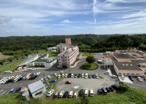 多層建物と駐車場を含む施設の上空からの全体像。周辺の緑地と道路が見える。